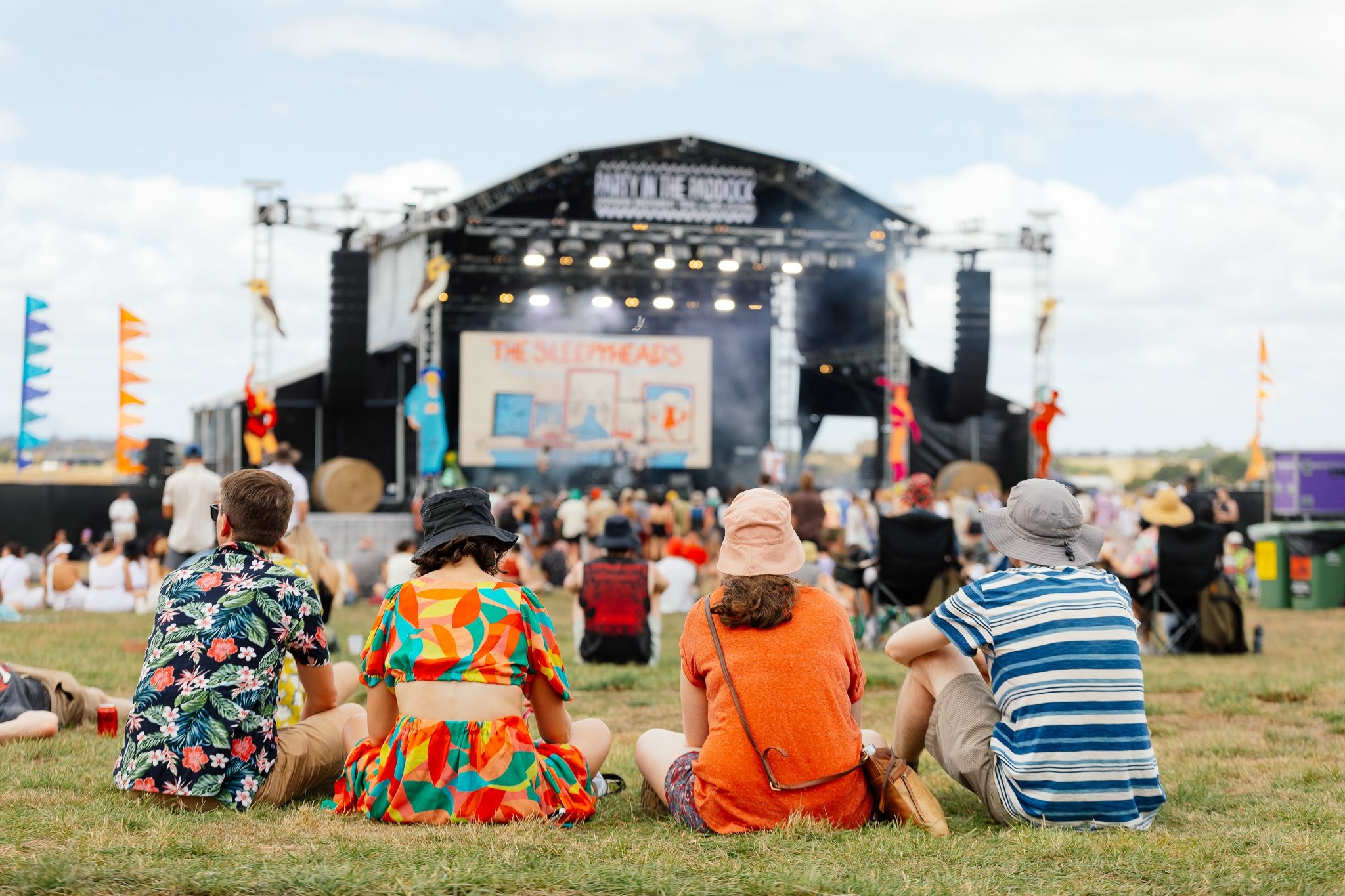 A group of friends sitting on the grass watching a band perform on a large outdoor festival stage, with colourful flags and a crowd of festival-goers visible across the sunny open field behind them.