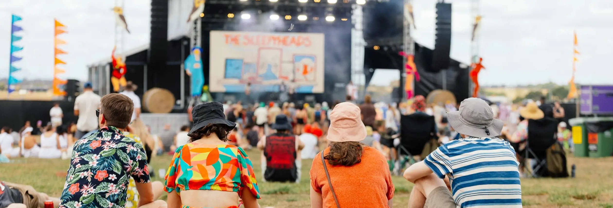 A group of friends sitting on the grass watching a band perform on a large outdoor festival stage, with colourful flags and a crowd of festival-goers visible across the sunny open field behind them.