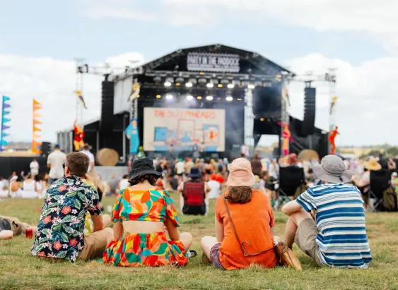 A group of friends sitting on the grass watching a band perform on a large outdoor festival stage, with colourful flags and a crowd of festival-goers visible across the sunny open field behind them.