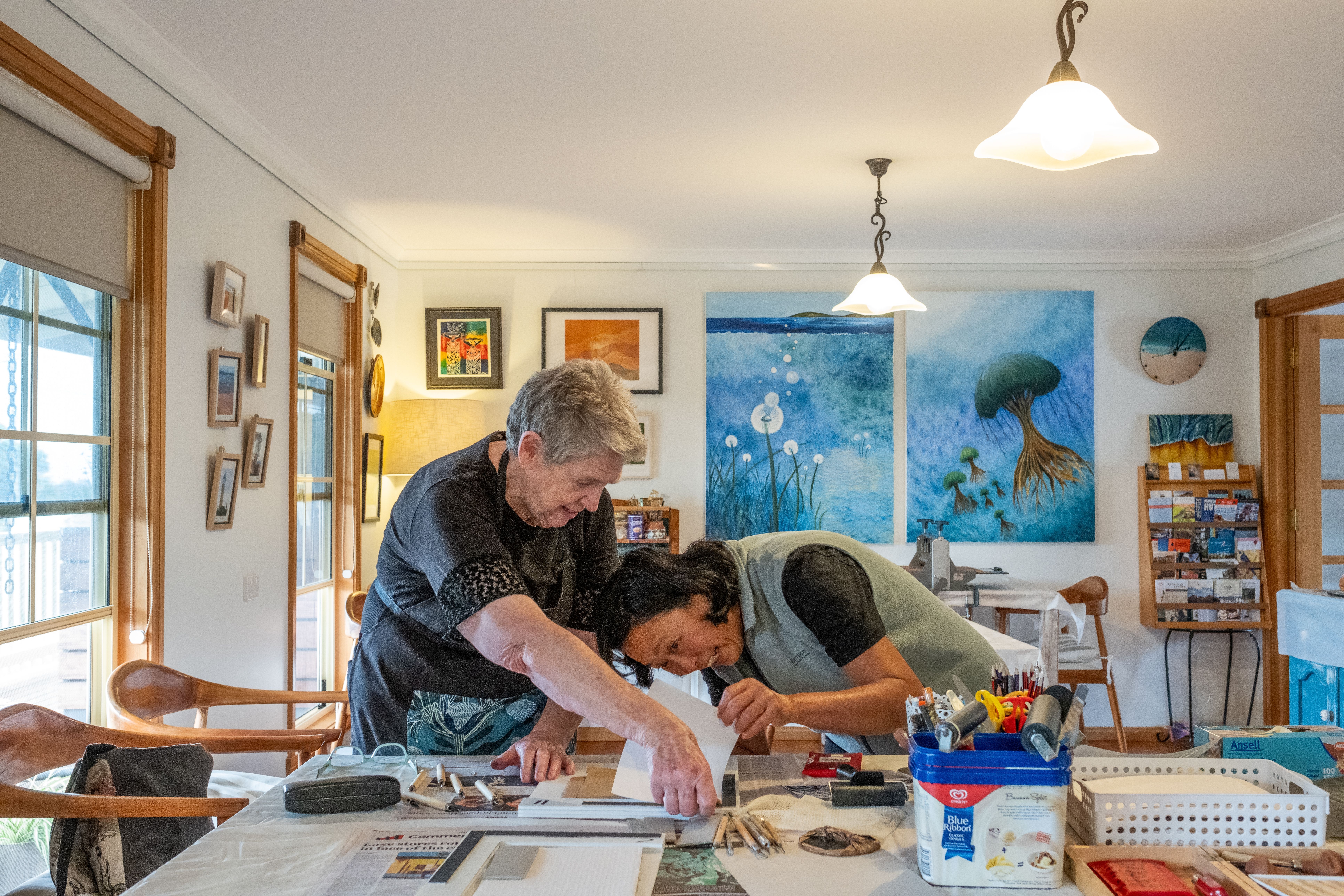 An artist leaning over a worktable to guide a student on a close detail of their work, in a bright home studio filled with colourful paintings, art supplies and natural light from large windows.