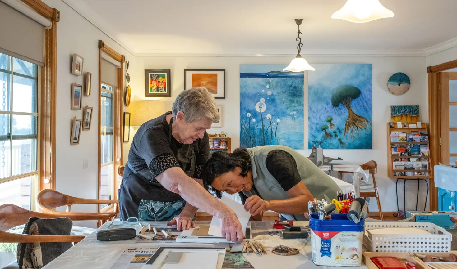 An artist leaning over a worktable to guide a student on a close detail of their work, in a bright home studio filled with colourful paintings, art supplies and natural light from large windows.