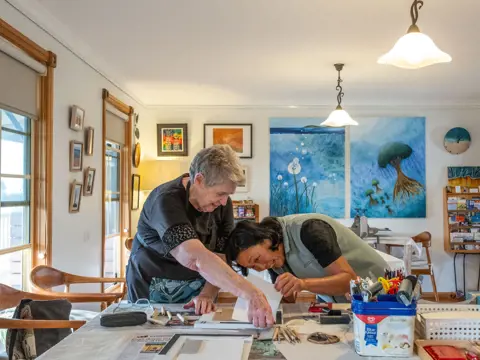 An artist leaning over a worktable to guide a student on a close detail of their work, in a bright home studio filled with colourful paintings, art supplies and natural light from large windows.