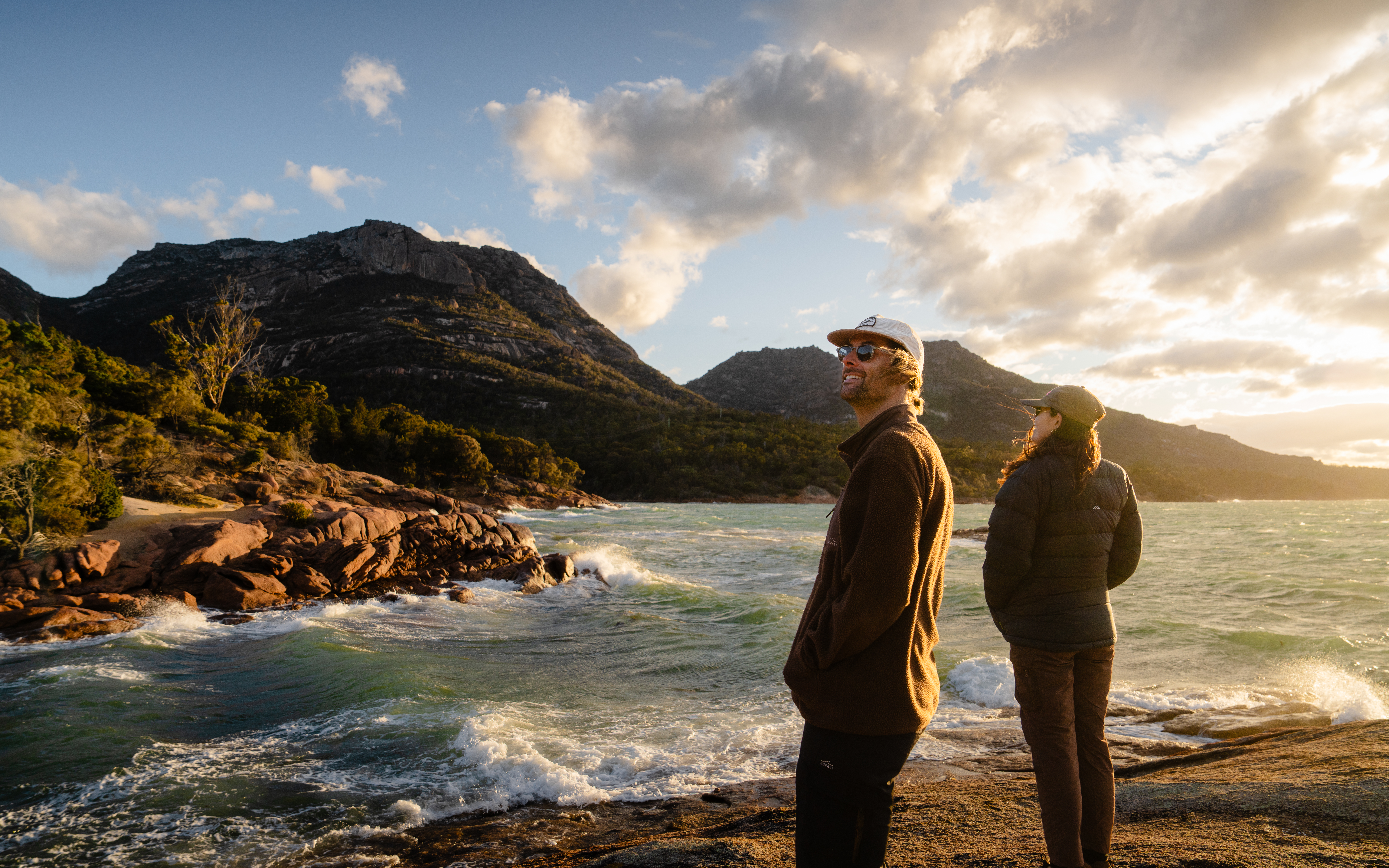 Two people standing on a rocky shoreline at sunset, looking out over a bay of choppy water with rugged granite headlands and dramatic clouds lit by warm golden light behind them.