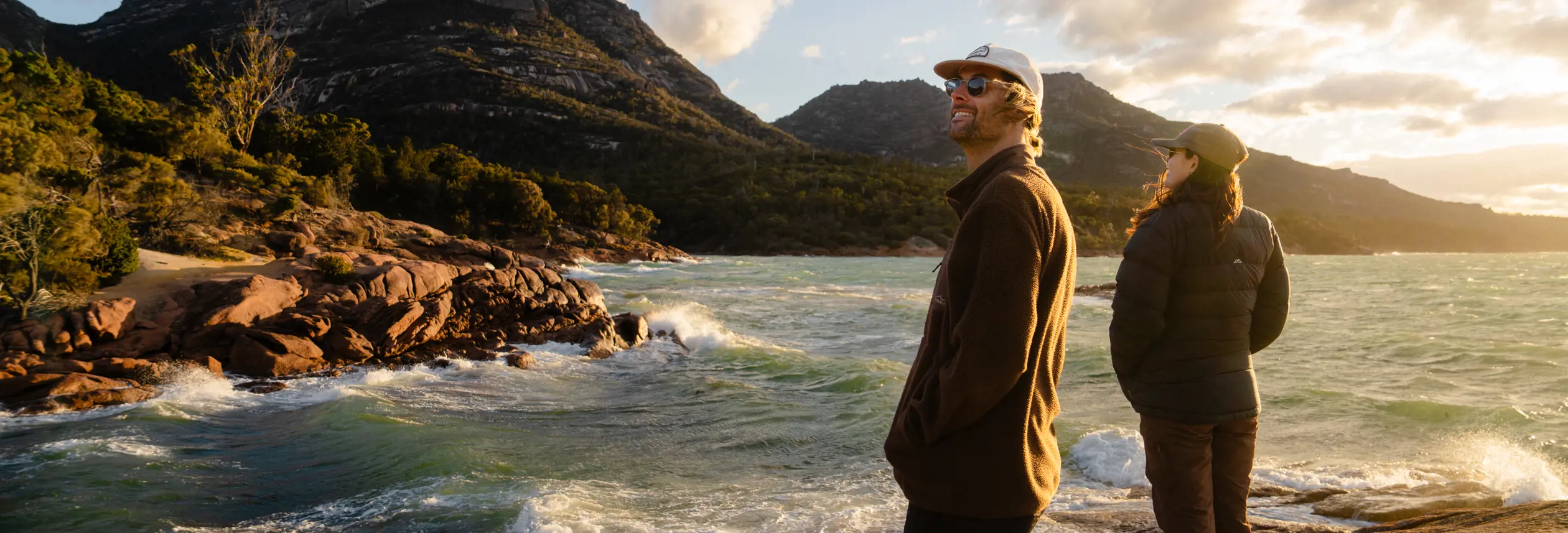 Two people standing on a rocky shoreline at sunset, looking out over a bay of choppy water with rugged granite headlands and dramatic clouds lit by warm golden light behind them.