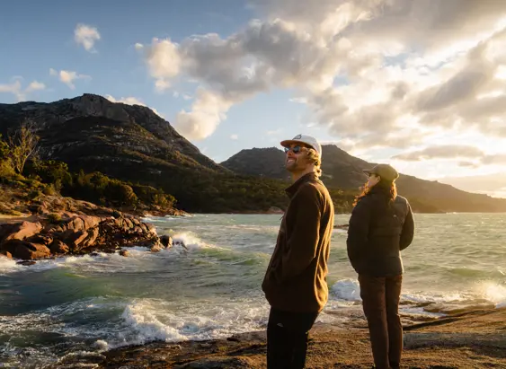 Two people standing on a rocky shoreline at sunset, looking out over a bay of choppy water with rugged granite headlands and dramatic clouds lit by warm golden light behind them.