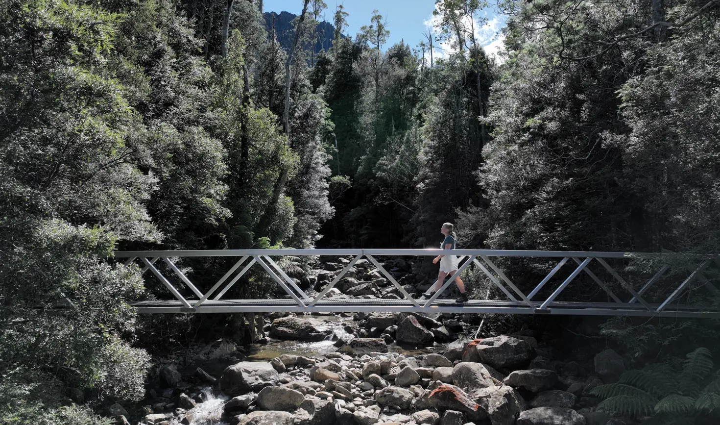 A walker crossing a narrow metal footbridge over a rocky mountain stream, surrounded by dense green forest and a glimpse of rocky peaks against a bright blue sky above.
