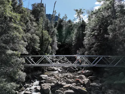 A walker crossing a narrow metal footbridge over a rocky mountain stream, surrounded by dense green forest and a glimpse of rocky peaks against a bright blue sky above.
