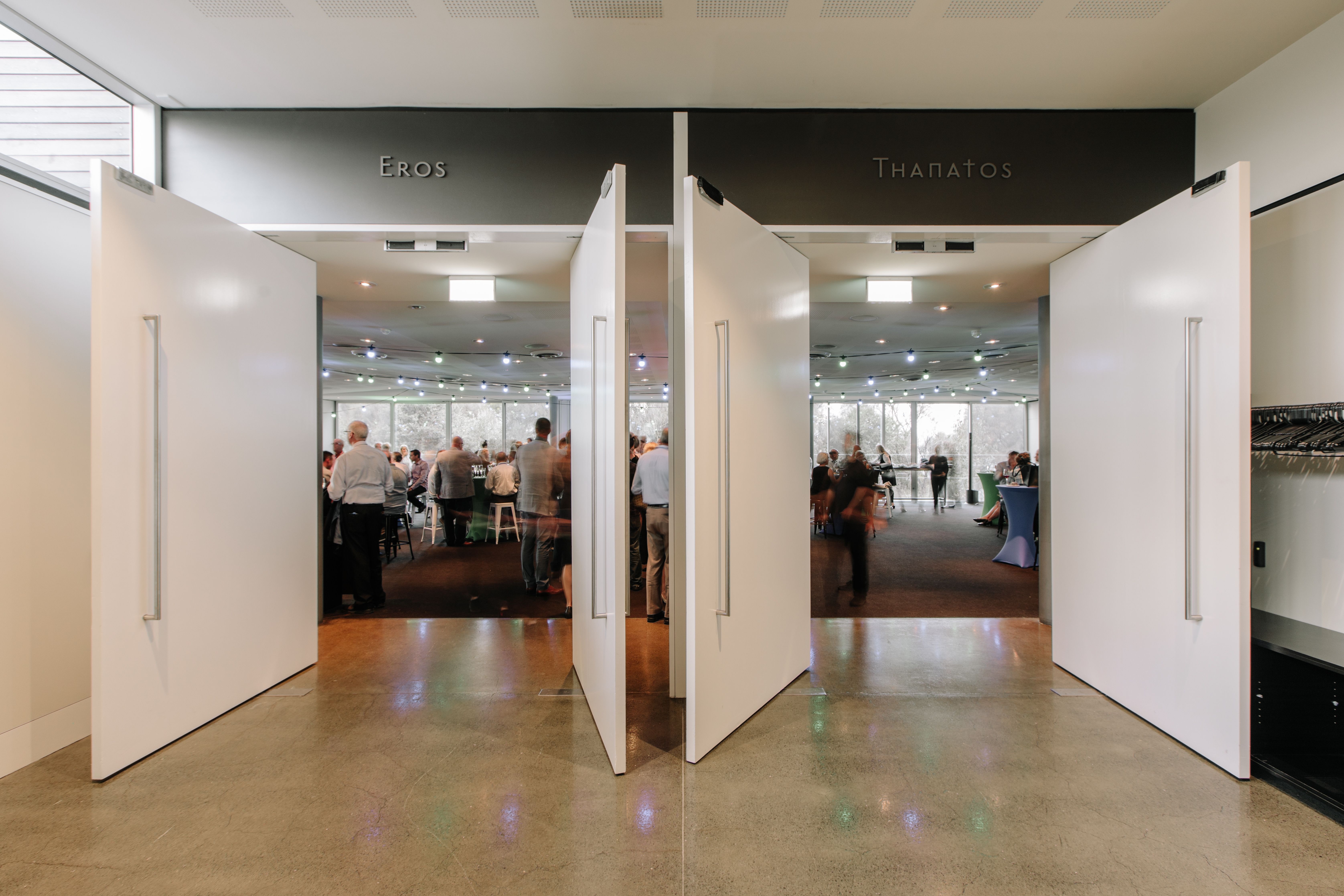 Two large white pivot doors opening into a bright, busy event or exhibition space, with people mingling inside and the words "Eros" and "Thanatos" displayed on the dark overhead panels above each entrance.