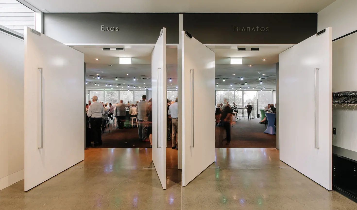 Two large white pivot doors opening into a bright, busy event or exhibition space, with people mingling inside and the words "Eros" and "Thanatos" displayed on the dark overhead panels above each entrance.