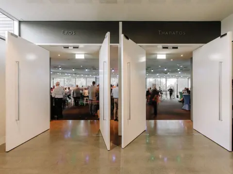 Two large white pivot doors opening into a bright, busy event or exhibition space, with people mingling inside and the words "Eros" and "Thanatos" displayed on the dark overhead panels above each entrance.
