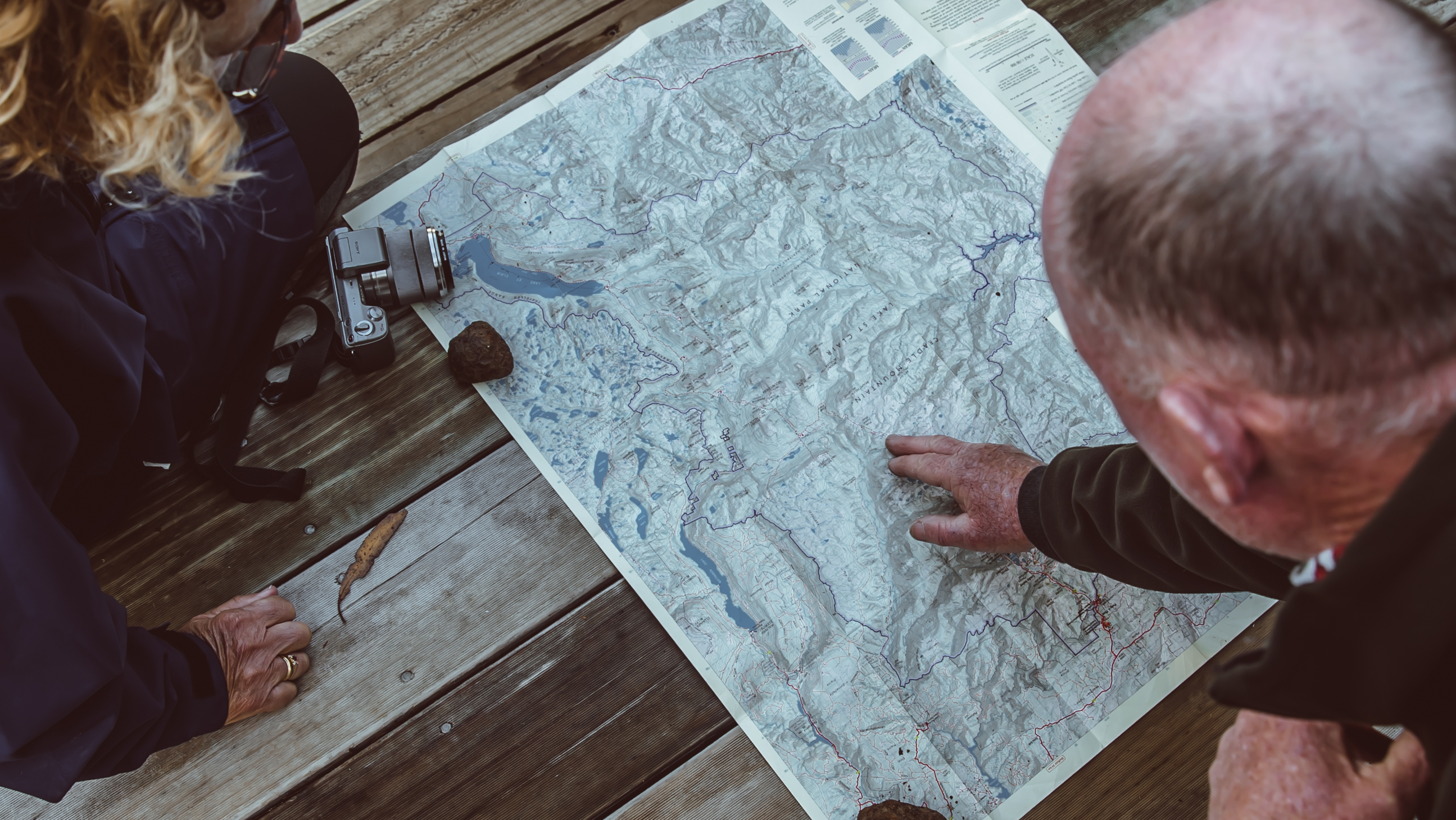 Two people leaning over a large topographic map spread on weathered timber decking, one pointing to a location as they plan their route.