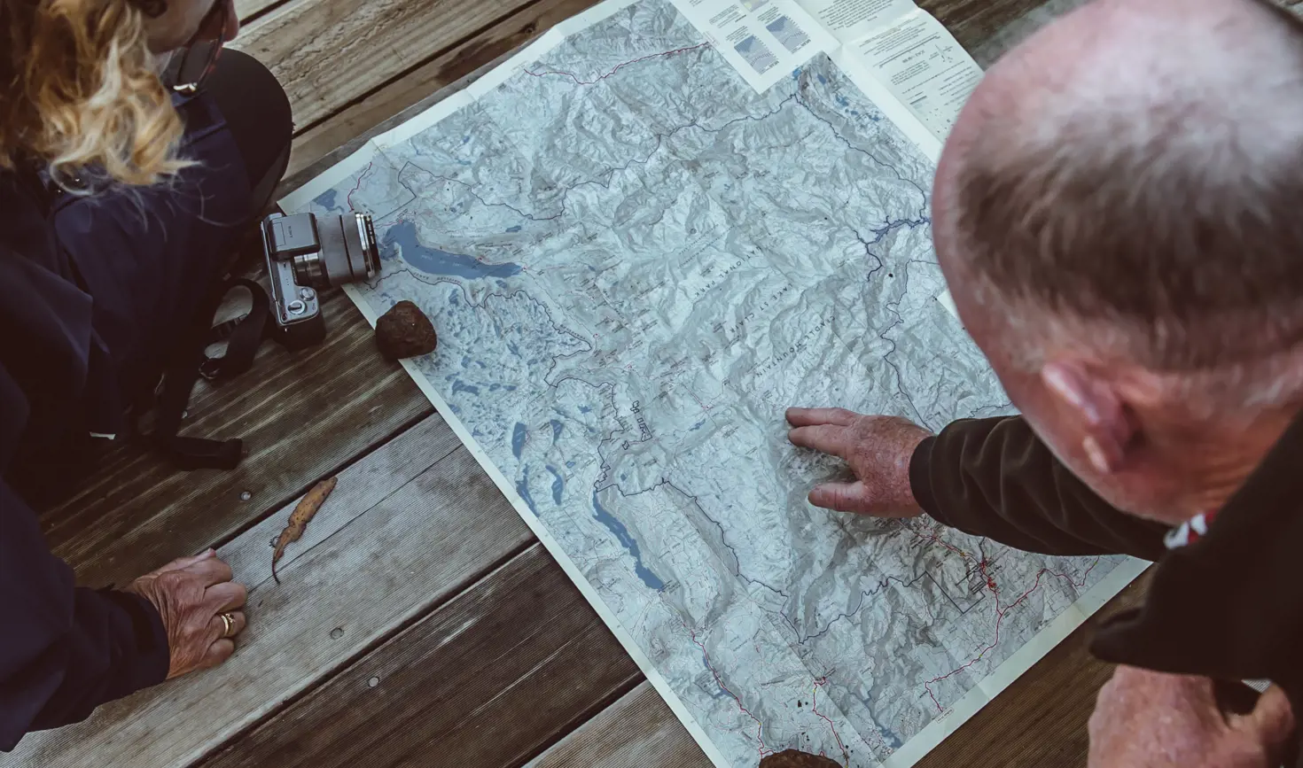 Two people leaning over a large topographic map spread on weathered timber decking, one pointing to a location as they plan their route.