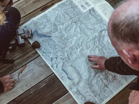 Two people leaning over a large topographic map spread on weathered timber decking, one pointing to a location as they plan their route.