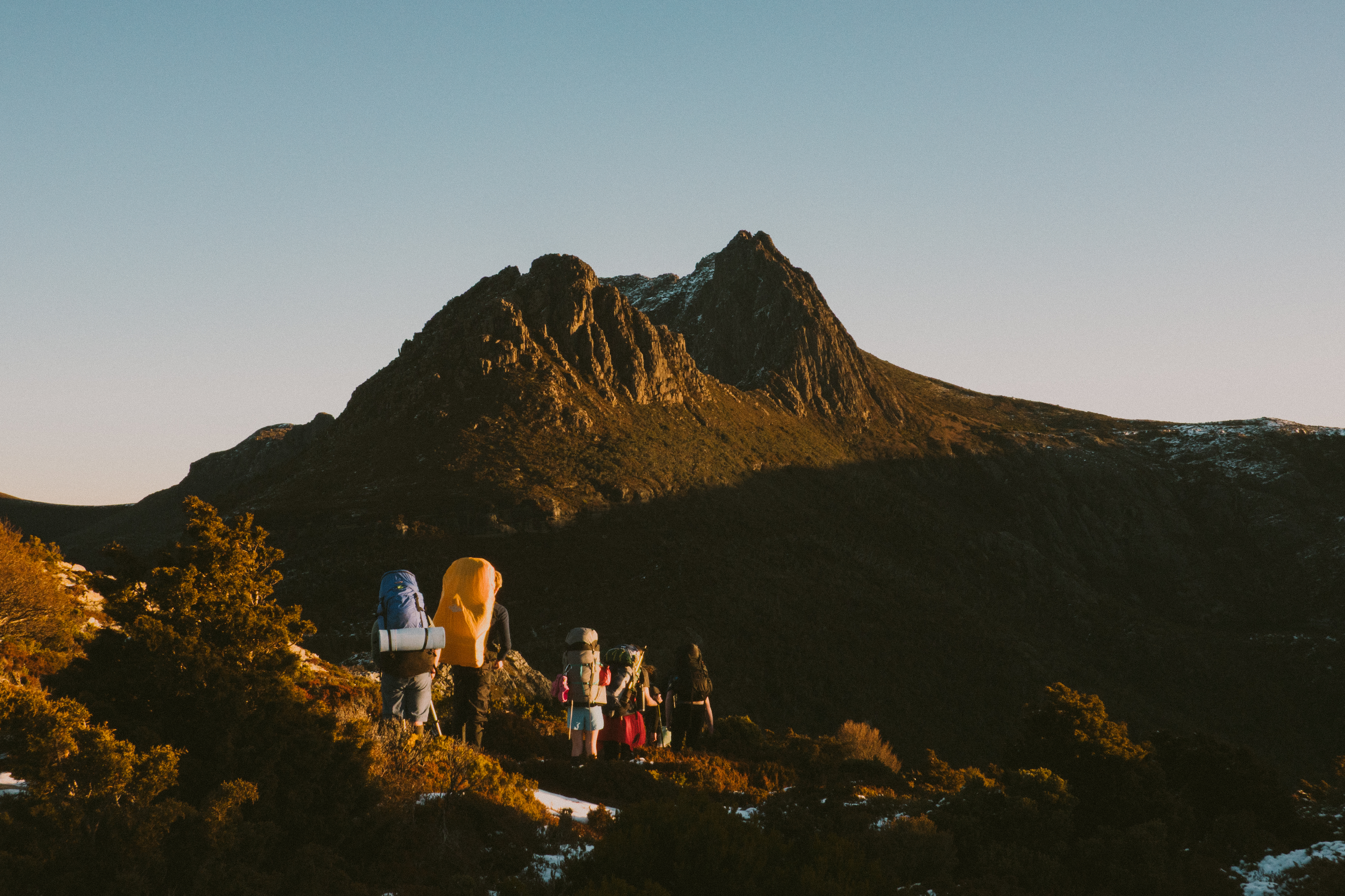 A group of hikers with large backpacks on a trail at golden hour, with a dramatic twin-peaked mountain rising against a clear sky ahead of them and patches of snow on the slopes below.