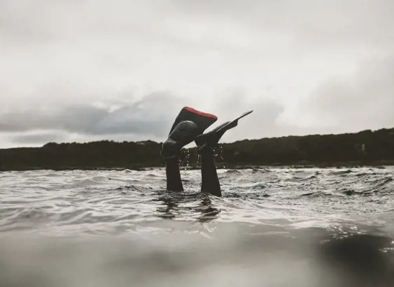 A diver in a wetsuit surface-diving into grey choppy water, their fins raised in the air as they descend, with a overcast sky and tree-lined shore visible in the background.