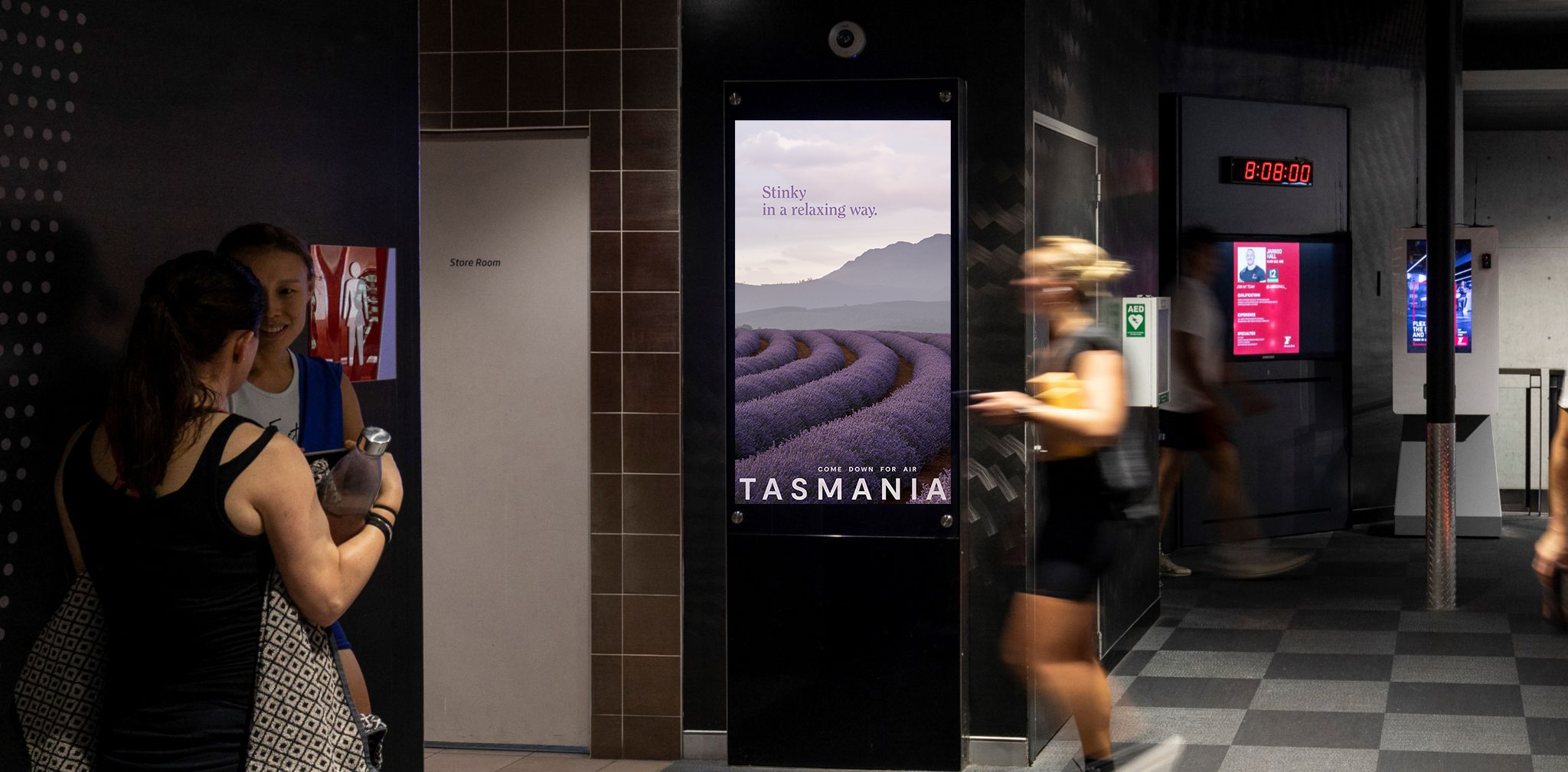 People walk past a poster in a subway. The poster shows purple lavender rows above the word: Tasmania.