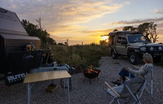 Lady in camp chair by an open fire and caravan with 4WD in background at sunset