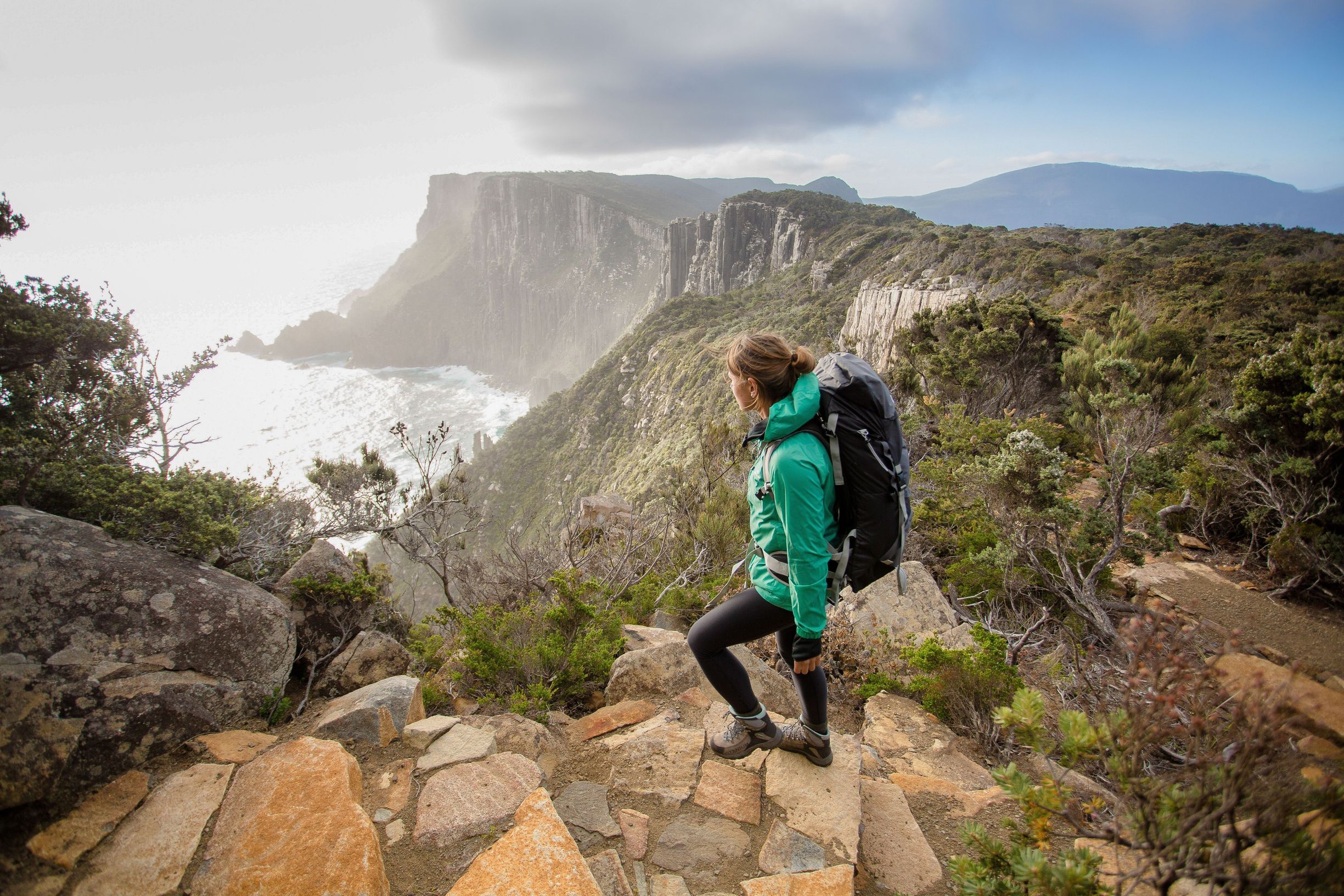A woman in a green coat and wearing a backpack, walks up rock steps on a rugged coastal track. She looks towards steep cliffs that rise from the sea.