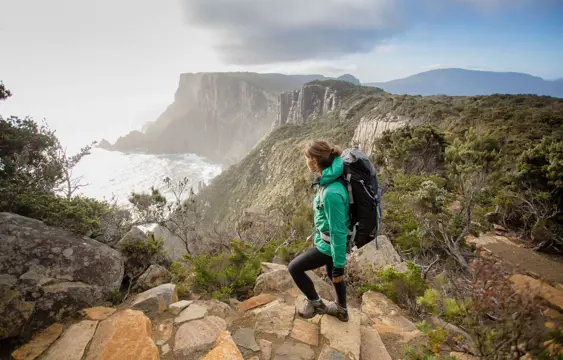 A woman in a green coat and wearing a backpack, walks up rock steps on a rugged coastal track. She looks towards steep cliffs that rise from the sea.