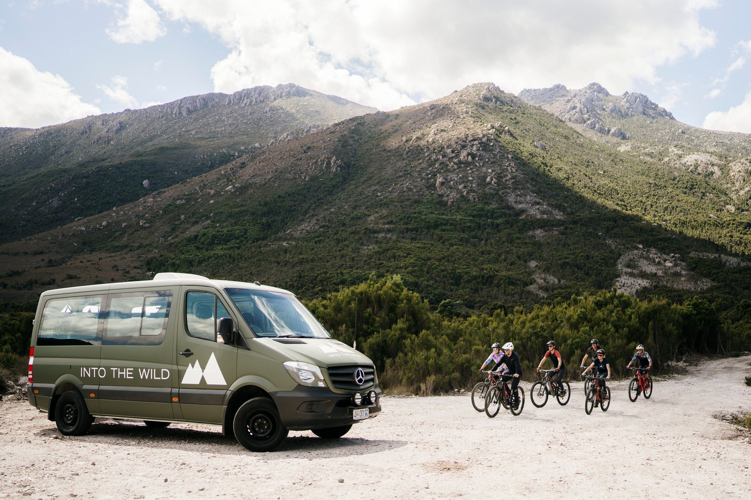 Six mountain bike riders cyle towards a khaki-coloured minivan on a cream gravel road as three rocky hills rise behind them.